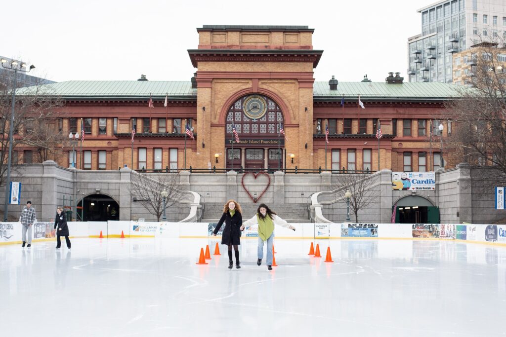 Two people ice skating at the Providence Rink in front of the historic Rhode Island Foundation building, showcasing a vibrant community winter activity with a heart-shaped decoration on the steps.